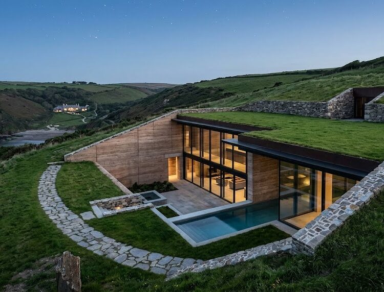 A stunning, minimalist luxury estate seamlessly carved into a grassy hillside, featuring an underground stone entrance and massive glass walls emerging from the landscape.